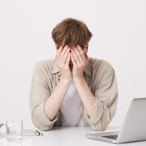 A person sits at a desk with their face in their hands, overwhelmed by tasks, yet determined to persevere like the teachings from the Stress Management and Resilience in the Workplace video program. An open laptop, surrounded by books and a notebook, hints at an ongoing battle for balance. A glass of water offers a moment of calm amidst chaos.