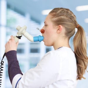 A woman in a clinical setting conducts the Spirometry Training Course using a spirometer, a medical device designed for assessing lung function. She holds the mouthpiece while standing in a brightly lit environment with a blurred background.